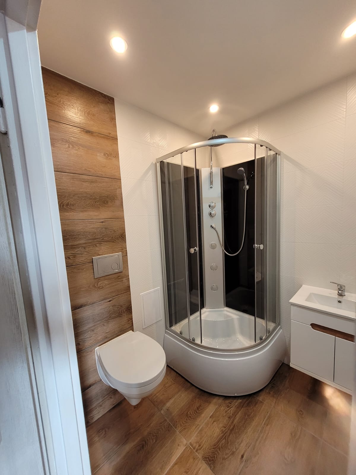 Nowoczesna łazienka po remoncie: kabina prysznicowa z hydromasażem, toaleta podtynkowa i umywalka z szafką. Płytki imitujące drewno i białe płytki z delikatnym wzorem.