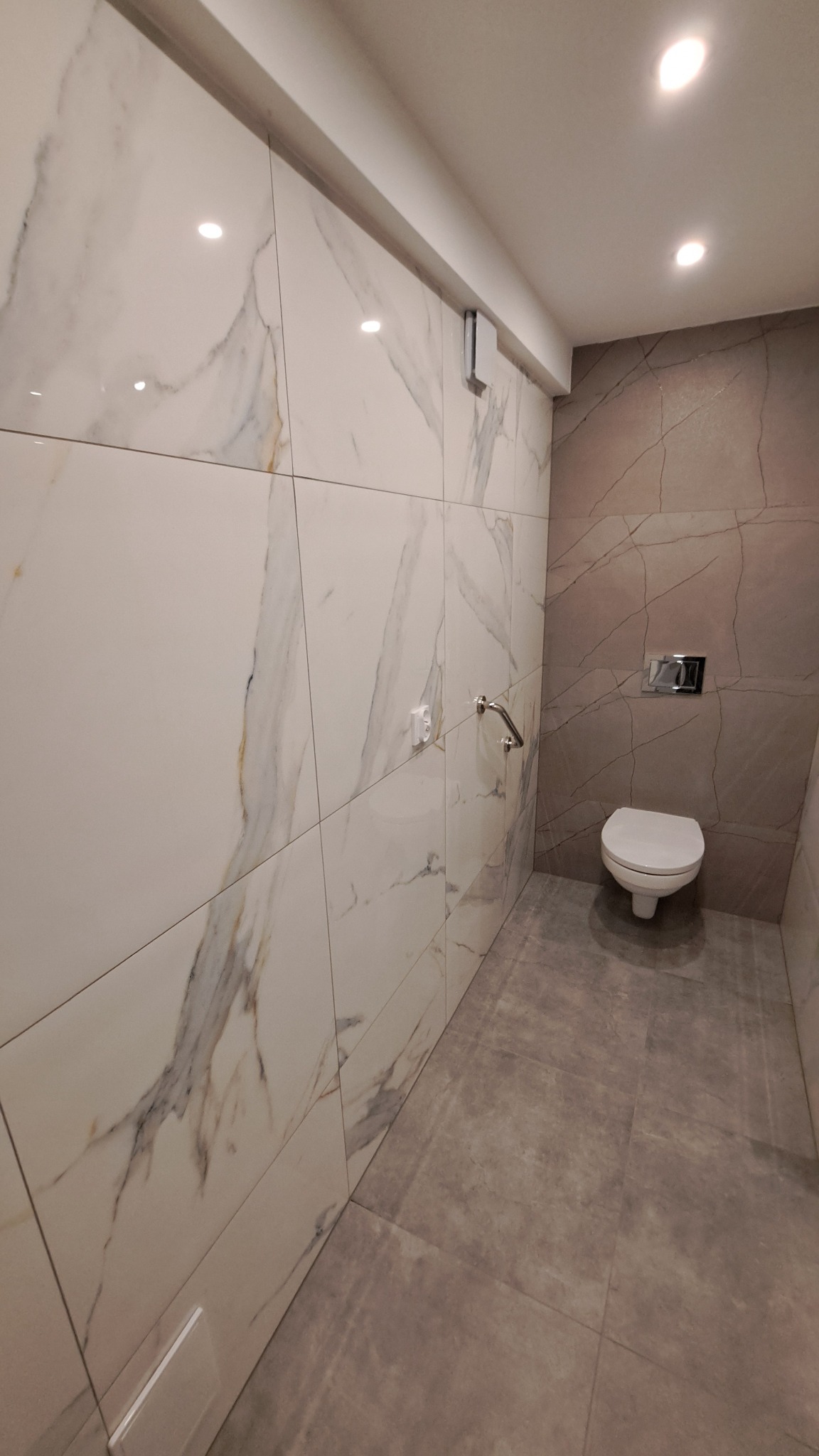 Nowoczesna łazienka z płytkami imitującymi marmur na ścianach i szarymi płytkami podłogowymi. Widoczna toaleta i chromowane uchwyty dla niepełnosprawnych.