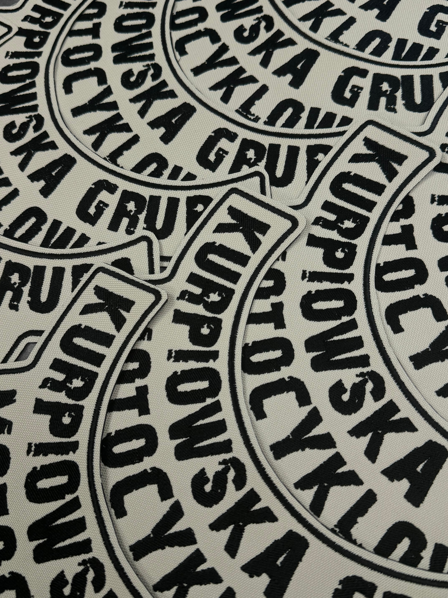 Zbliżenie na naszywki z czarnym tekstem 'KURPIOWSKA GRUPA KOLARSKA' na jasnym tle, detale szwów i faktura materiału widoczne.