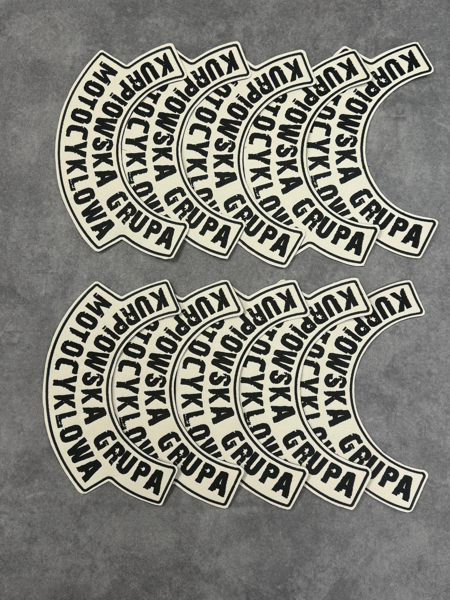 Partia naszywek z napisem 'Kurpiowska Motocyklowa Grupa', leżących na szarym tle, z bliska widoczna struktura materiału.