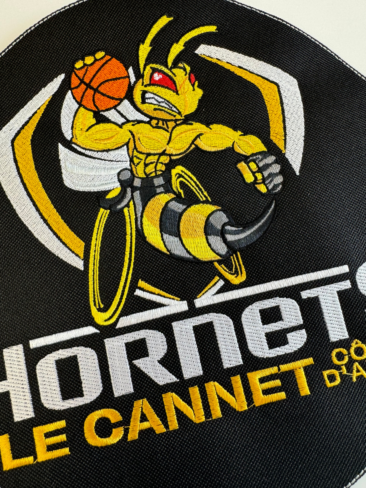 Czarna naszywka z haftowanym wizerunkiem agresywnej osy grającej w koszykówkę, z napisem 'HORNETS LE CANNET', wykonana techniką haftu maszynowego z dbałością o detale.