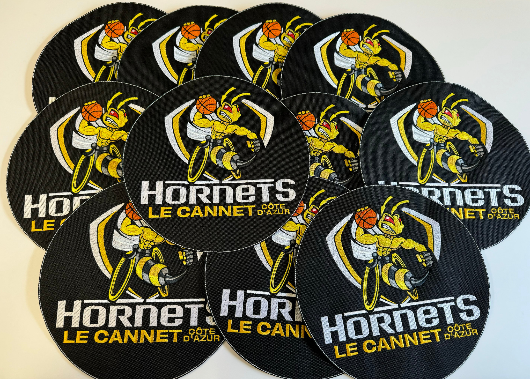 Seria okrągłych naszywek z wyhaftowanym logo drużyny sportowej Hornets Le Cannet Côte d'Azur, przedstawiającym osę trzymającą piłkę do koszykówki, ułożonych na białym tle.