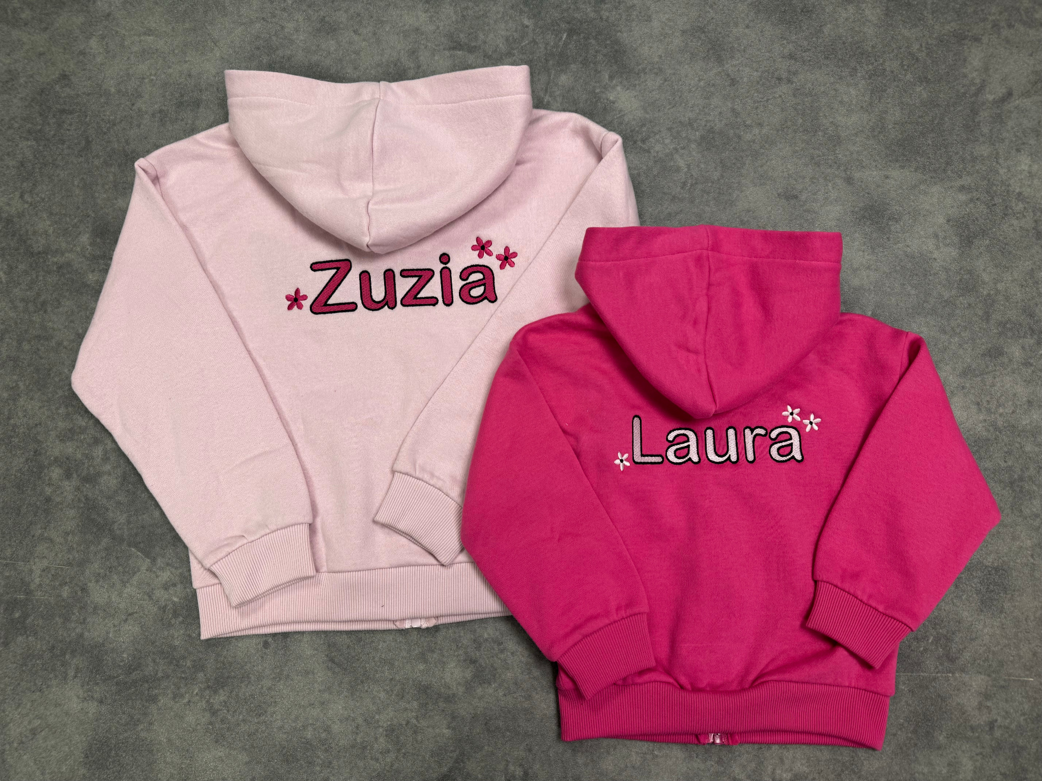 Dwa rozłożone, dziecięce, zapinane na zamek bluzy z kapturem: jasnoróżowa z haftowanym imieniem 'Zuzia' i małą kwiatową dekoracją oraz fuksjowa z haftowanym imieniem 'Laura' i kwiatową dekoracją...