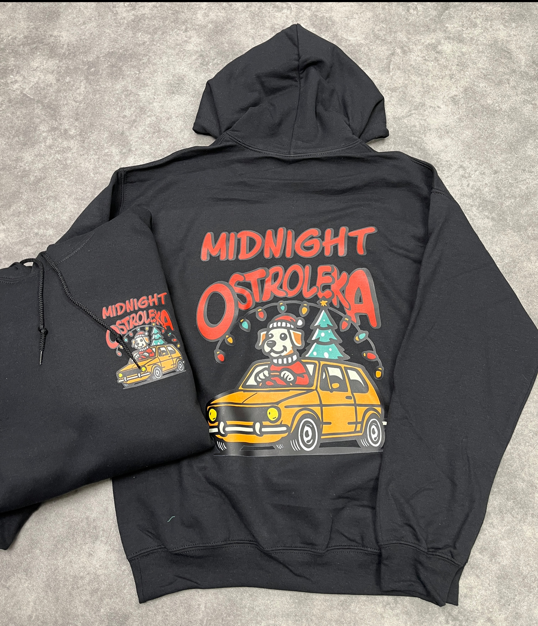 Dwa czarne swetry z nadrukiem w Ostrołęce: pies w czapce Mikołaja prowadzący żółty samochód z choinką na dachu, otoczony lampkami, z napisem 'Midnight Ostrołęka'.