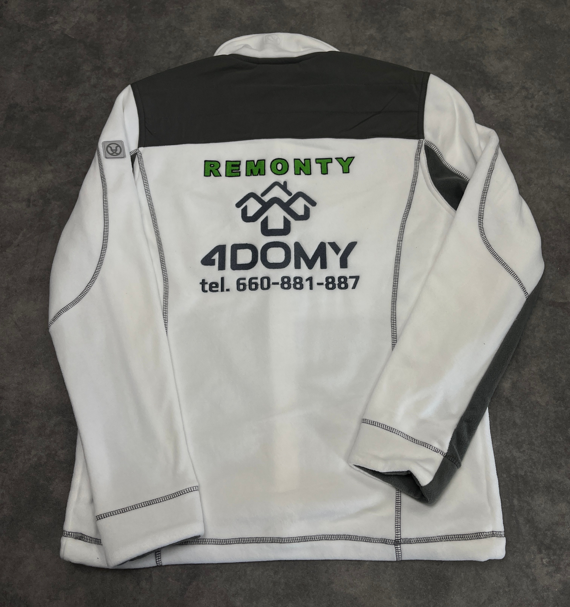 Biała bluza polarowa z szarymi wstawkami i nadrukiem firmy remontowej 'Remonty 4Domy' wraz z numerem telefonu, leżąca na szarym tle.