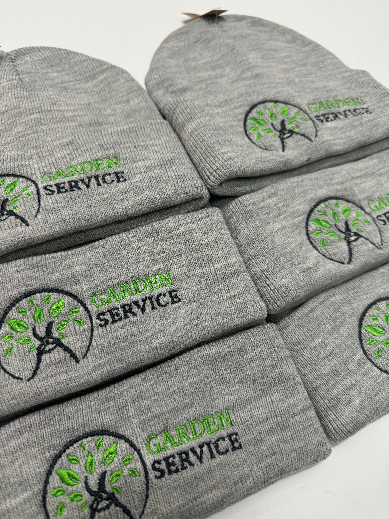 Stos szarych czapek zimowych z wyhaftowanym logo 'Garden Service' w kolorze zielonym i czarnym, ułożonych na białym tle.