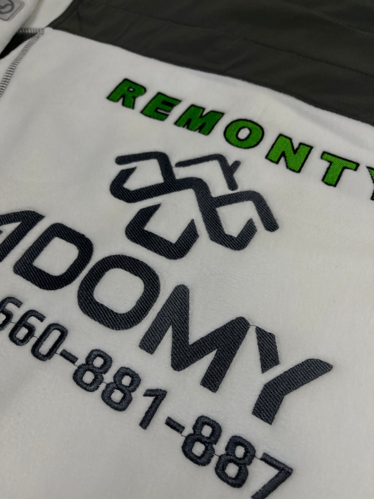 Biała bluza polarowa z wyhaftowanym zielonym napisem 'REMONT' i szarym napisem 'ADOMY' oraz numerem telefonu w szarej nici.
