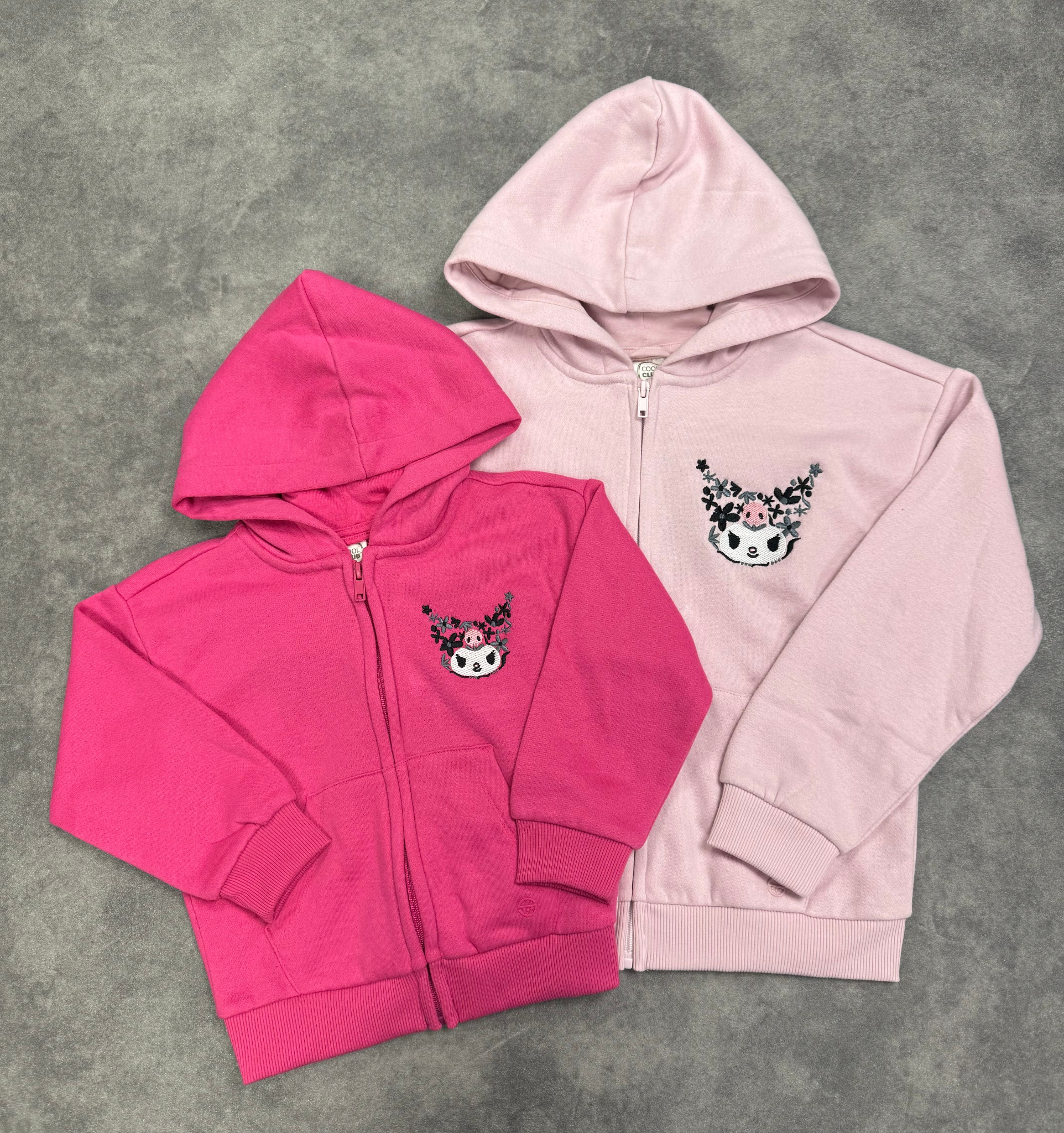 Dwie rozpięte bluzy z kapturem, jedna różowa, druga fuksjowa, obie z haftowanym motywem postaci z czarnymi kwiatami na piersi, leżące na szarym tle.