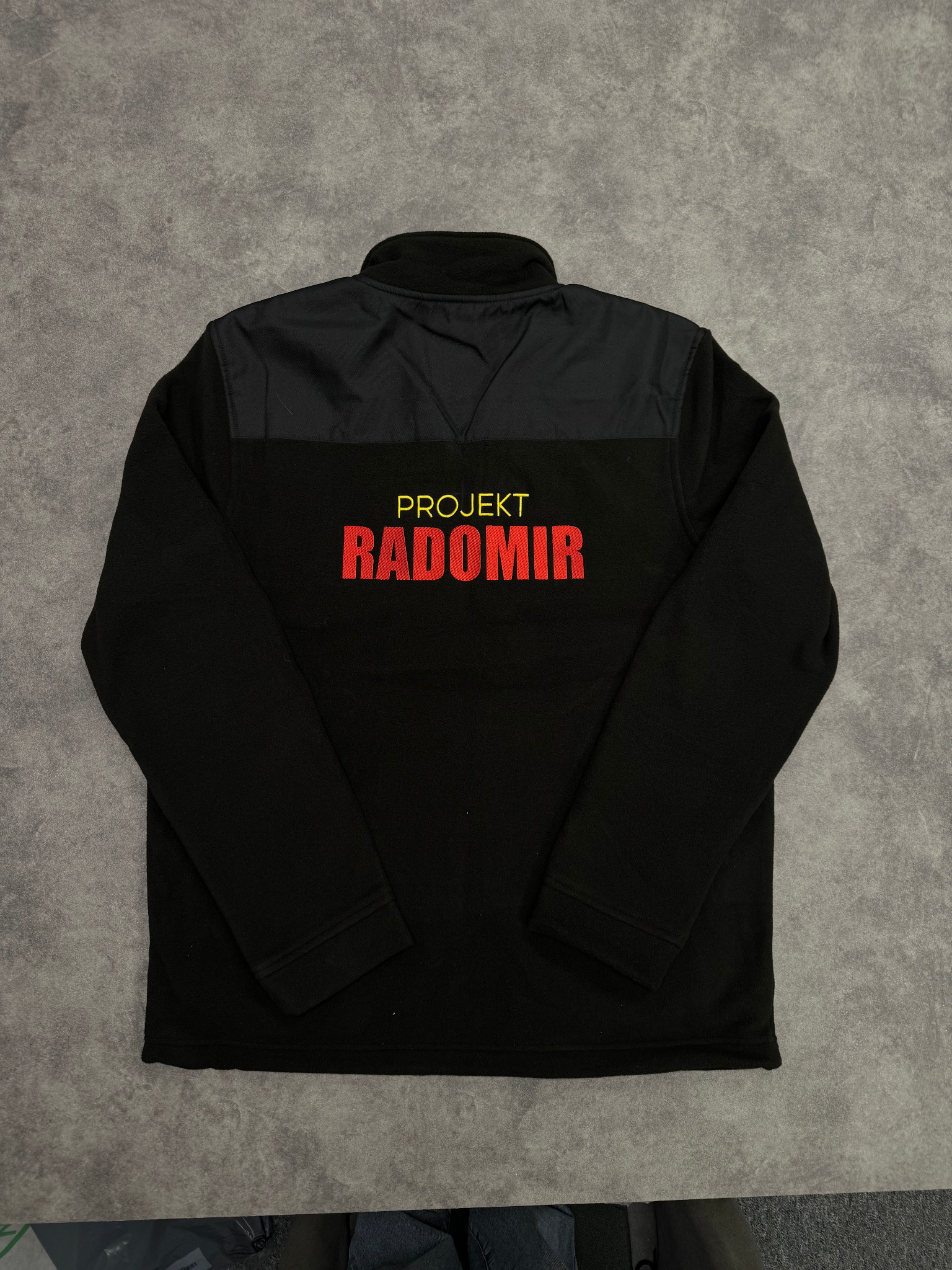 Czarna bluza polarowa z krótkim zamkiem, z nadrukiem 'Projekt Radomir' w kolorze czerwonym, leżąca na szarym tle.