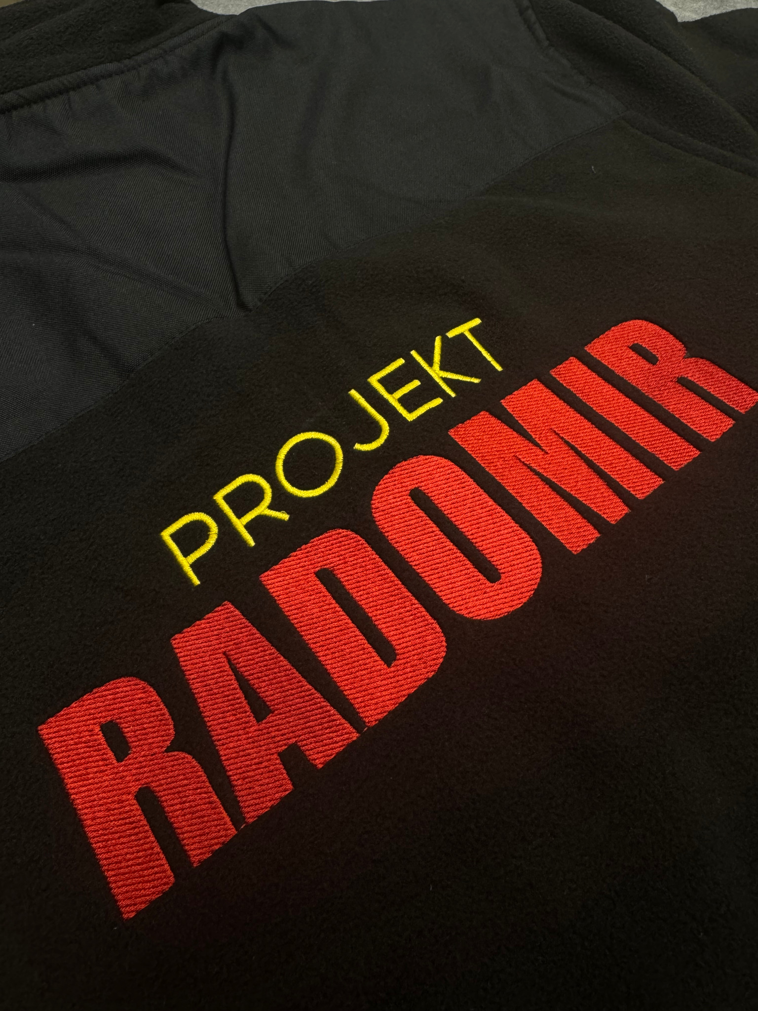 Czarna bluza z kontrastowym górnym panelem i wyhaftowanym napisem 'PROJEKT RADOMIR' w kolorach żółtym i czerwonym, widoczne detale ściegów.