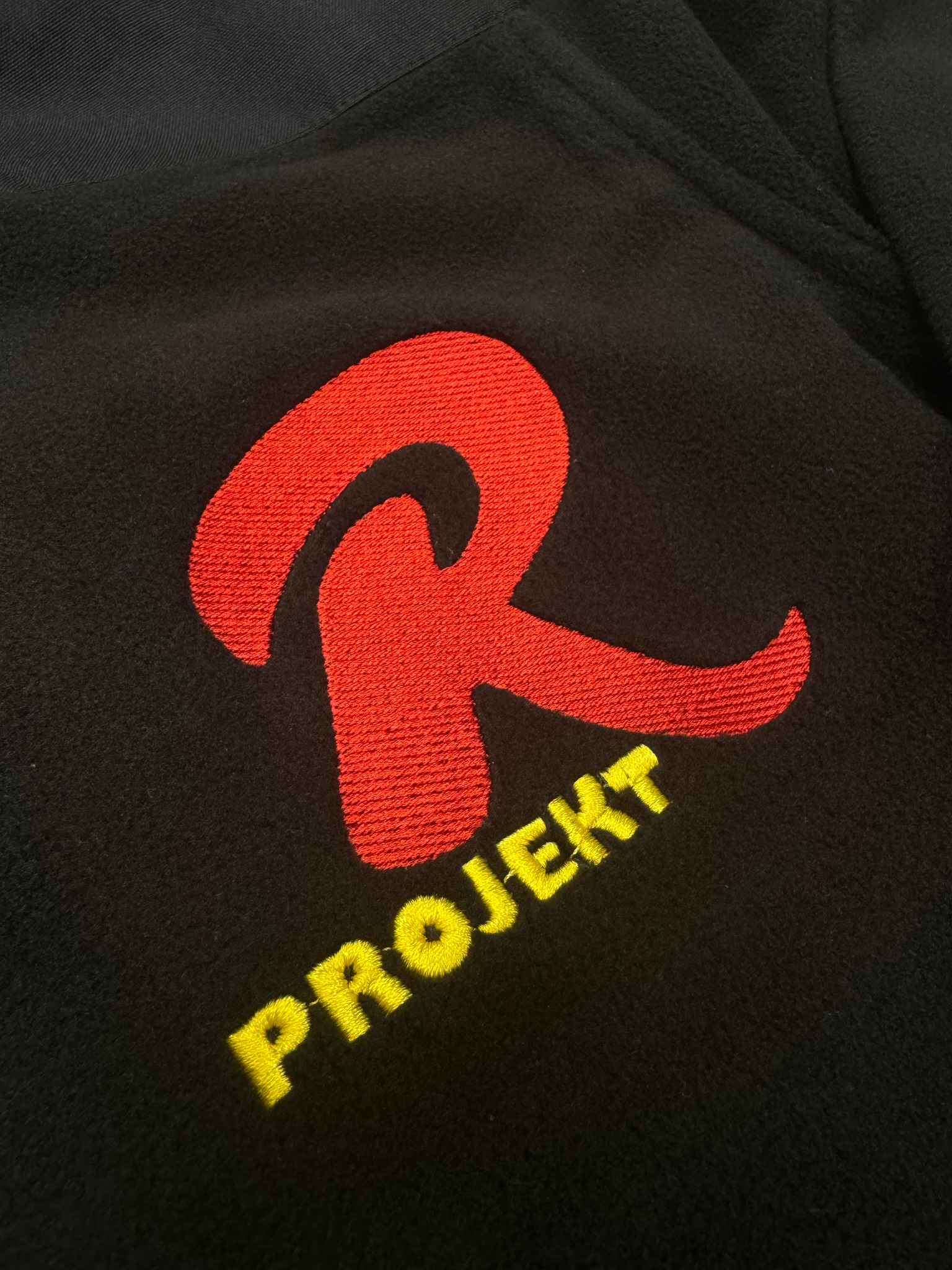 Czarna bluza polarowa z wyhaftowanym czerwonym logo litery 'R' i żółtym napisem 'PROJEKT' poniżej.