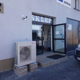 Zewnętrzna jednostka klimatyzacji marki Immergas, zainstalowana przed wejściem do sklepu z otwartymi drzwiami, na betonowych podstawach, z widocznymi elementami wentylacyjnymi na ścianie budynku.
