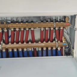 Rozdzielacz ogrzewania podłogowego w metalowej szafce, z widocznymi rurami w kolorze czerwonym i niebieskim, zaworami oraz numeracją obwodów. Na drzwiczkach szafki widoczne odręczne notatki. Widok...