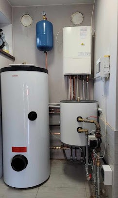 Zestaw urządzeń grzewczych w kotłowni: dwa białe zbiorniki, niebieski zbiornik wyrównawczy, piec wiszący z miedzianymi rurami, instalacja połączona mosiężnymi kształtkami, widoczne ślady po...