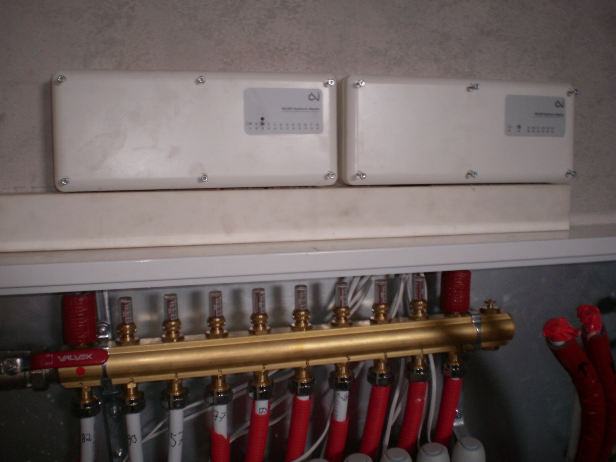 Rozdzielacz ogrzewania podłogowego z rurami w izolacji, zamontowany w szafce pod dwoma białymi sterownikami Wilo Hydronic Master, widoczne numery na rurach.