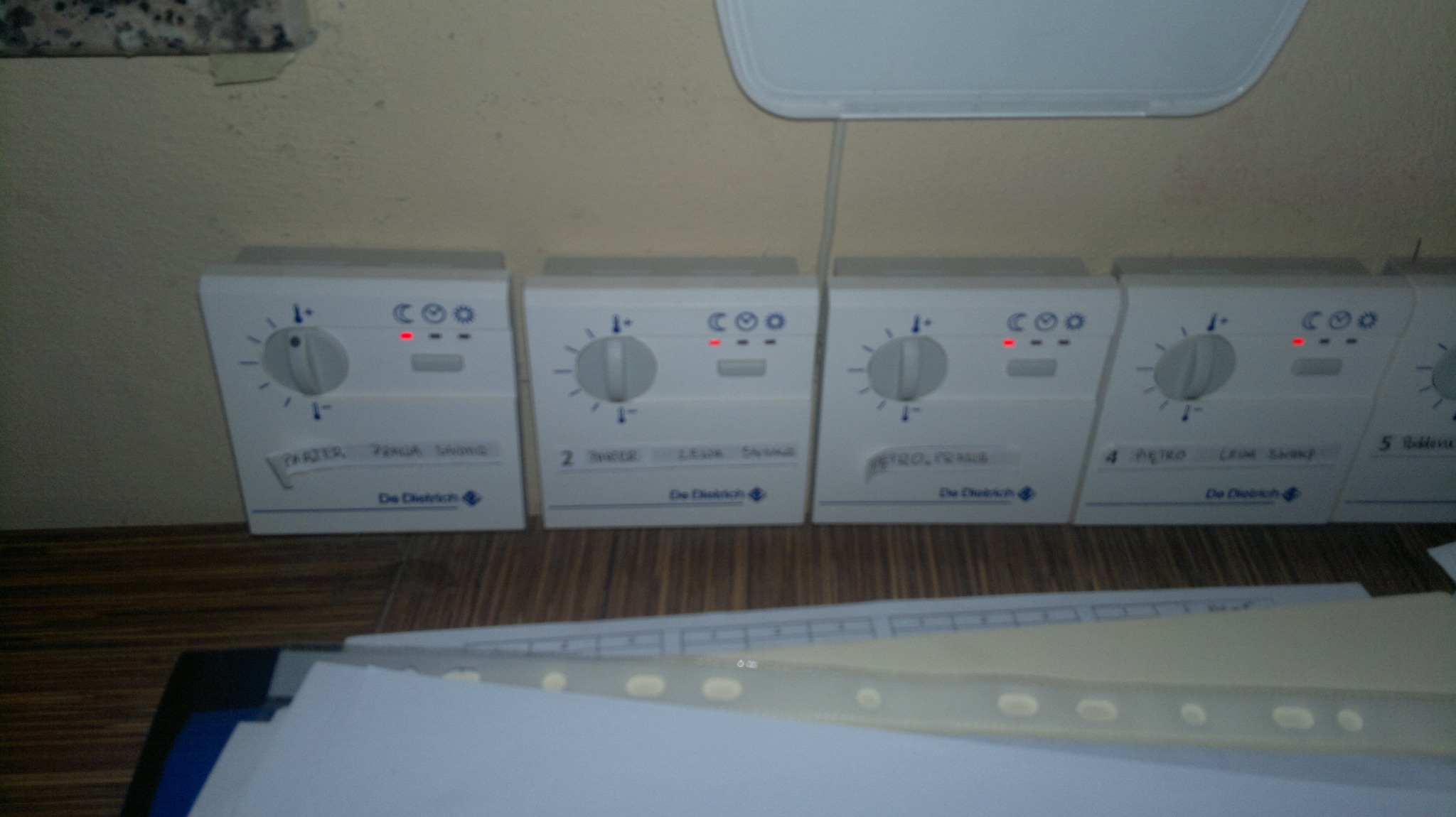 Szereg białych regulatorów temperatury De Dietrich z czerwonymi diodami LED, umieszczonych na ścianie nad podłogą z paneli. Widoczne etykiety z nazwami pomieszczeń: Parter, 2, 4, 5. Na pierwszym...