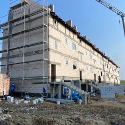 Tom&Tom - Wielopiętrowy budynek w trakcie budowy, z widocznymi rusztowaniami, częściowo otynkowaną elewacją, niezamontowanymi oknami i pracownikami na placu budowy, z dźwigiem w tle.