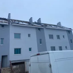 Trzysegmentowy budynek mieszkalny z szarą elewacją, widoczne okna zabezpieczone folią, dach pokryty śniegiem, na pierwszym planie fragment białego samochodu dostawczego.