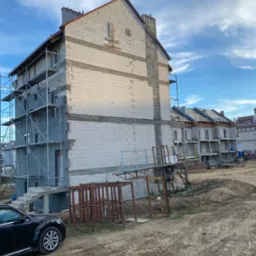 Budynek w trakcie budowy elewacji z widocznymi rusztowaniami, pustakami i elementami konstrukcyjnymi, na tle błękitnego nieba z chmurami.