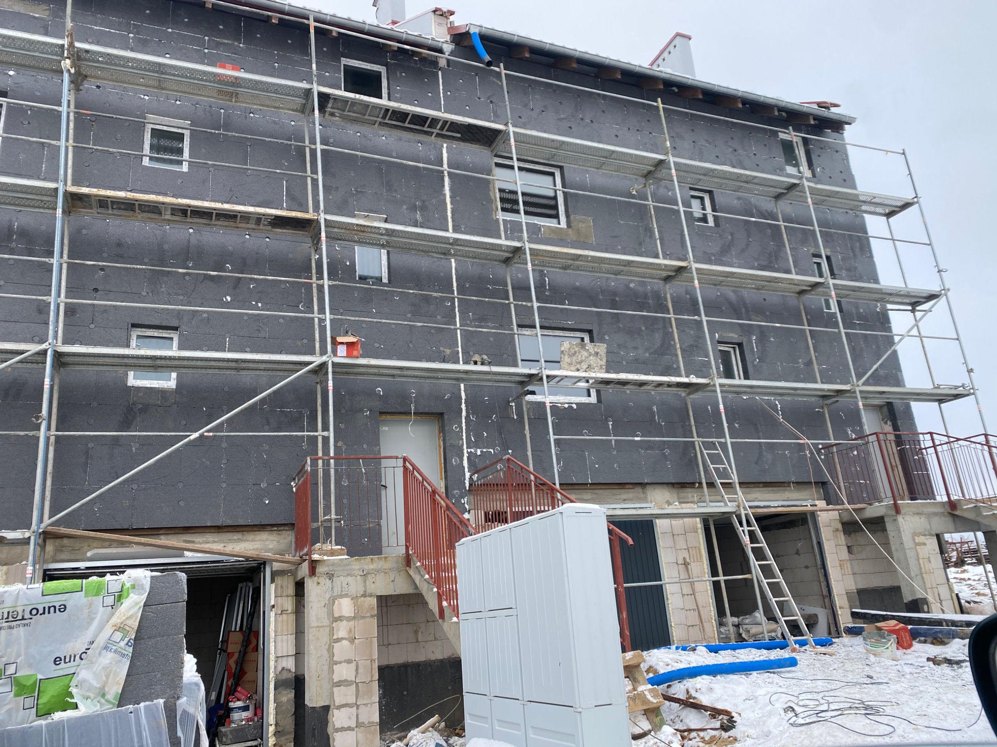 Budynek wielorodzinny w trakcie ocieplania, widoczne rusztowania, czarna warstwa izolacyjna na elewacji, materiały budowlane oraz elementy wykończeniowe na pierwszym planie, zimowa sceneria.