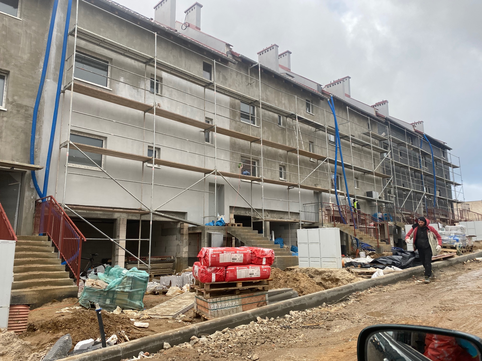 Budynek wielorodzinny w trakcie ocieplania, widoczne rusztowania, materiały izolacyjne Rockwool na paletach, nieotynkowana elewacja i niebieskie rury wentylacyjne.