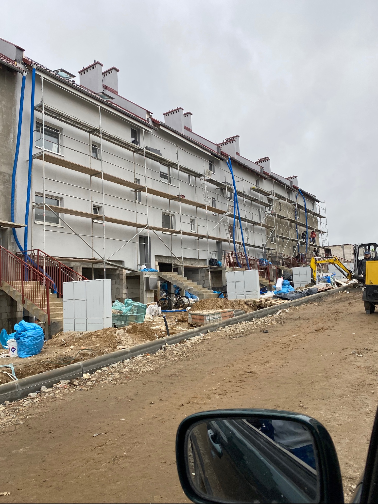 Budynek wielorodzinny w trakcie ocieplania, widoczne rusztowania, niebieskie rury wentylacyjne, mini koparka i robotnicy na rusztowaniach, teren budowy w nieładzie, widok z samochodu.