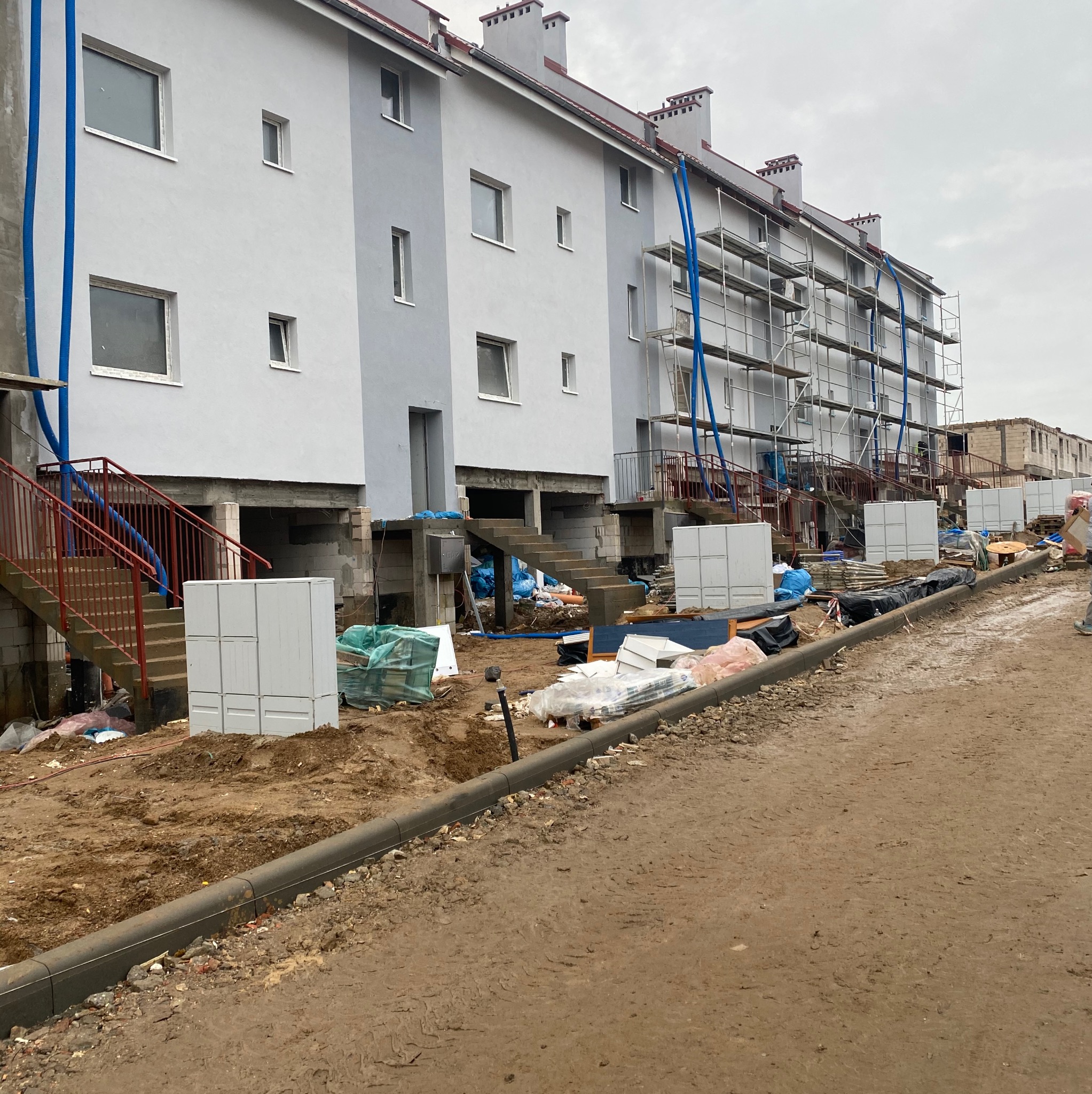 Wielorodzinny budynek w trakcie ocieplania elewacji, widoczne rusztowania, niebieskie rury do transportu materiałów, przygotowane białe płyty izolacyjne i teren budowy.