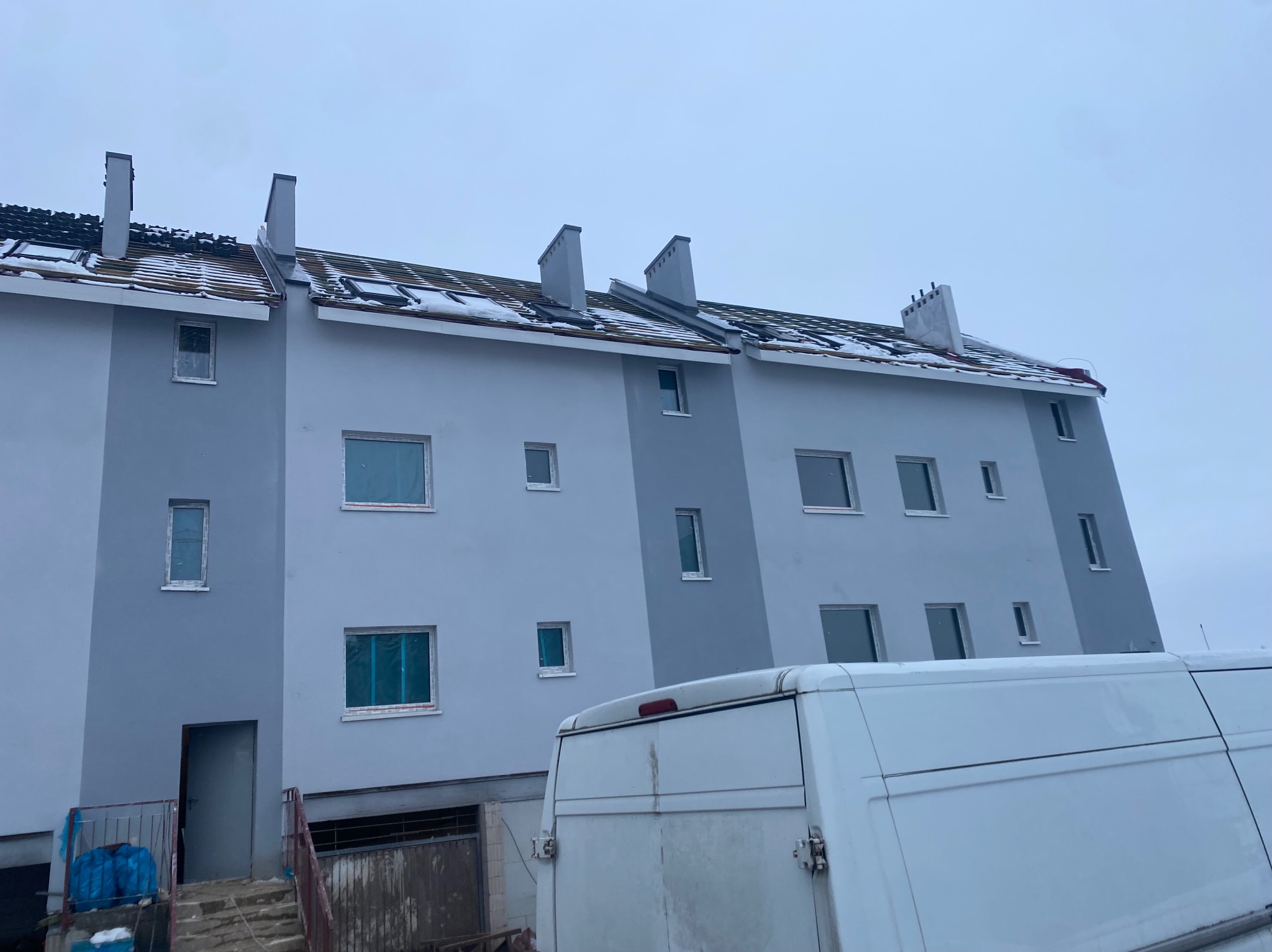 Trzysegmentowy budynek mieszkalny z szarą elewacją, widoczne okna zabezpieczone folią, dach pokryty śniegiem, na pierwszym planie fragment białego samochodu dostawczego.