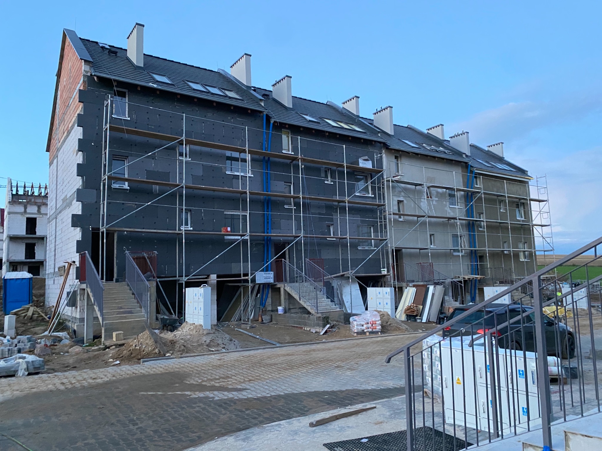 Budynek wielorodzinny w trakcie ocieplania, widoczne rusztowania, styropian grafitowy na elewacji, fragmenty elewacji w kolorze szarym, nieukończona elewacja z cegły, plac budowy i materiały...