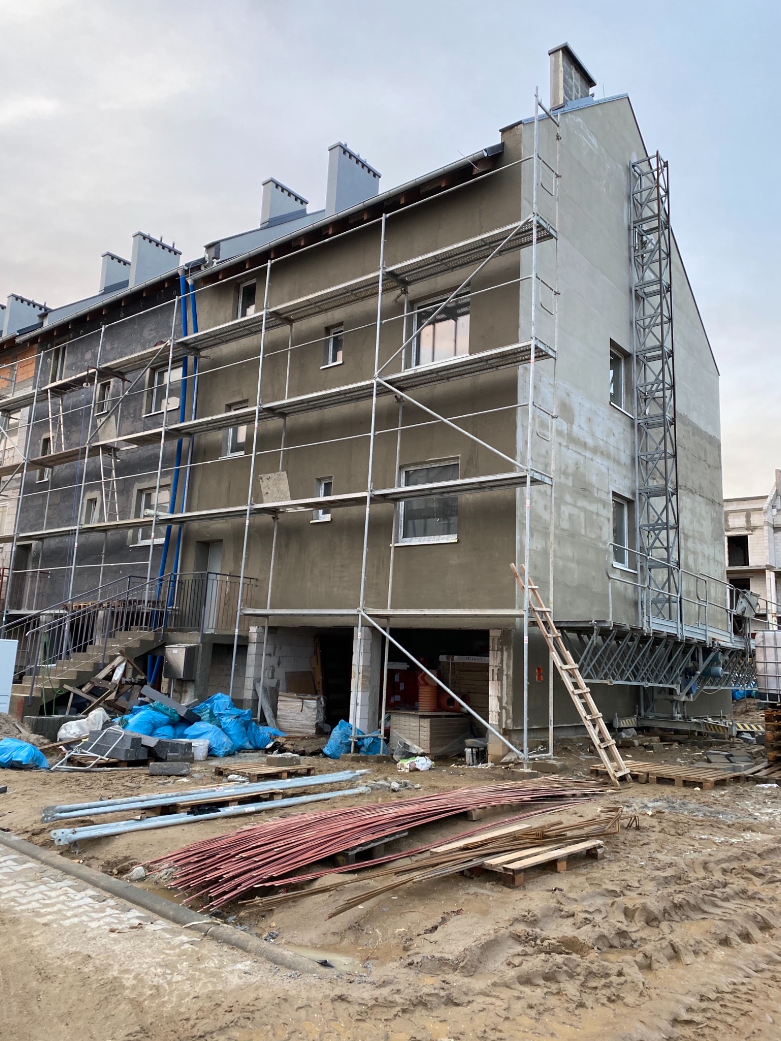 Budynek wielorodzinny w trakcie ocieplania, widoczne rusztowania, worki z materiałem izolacyjnym i stosy prętów zbrojeniowych na błotnistym terenie.