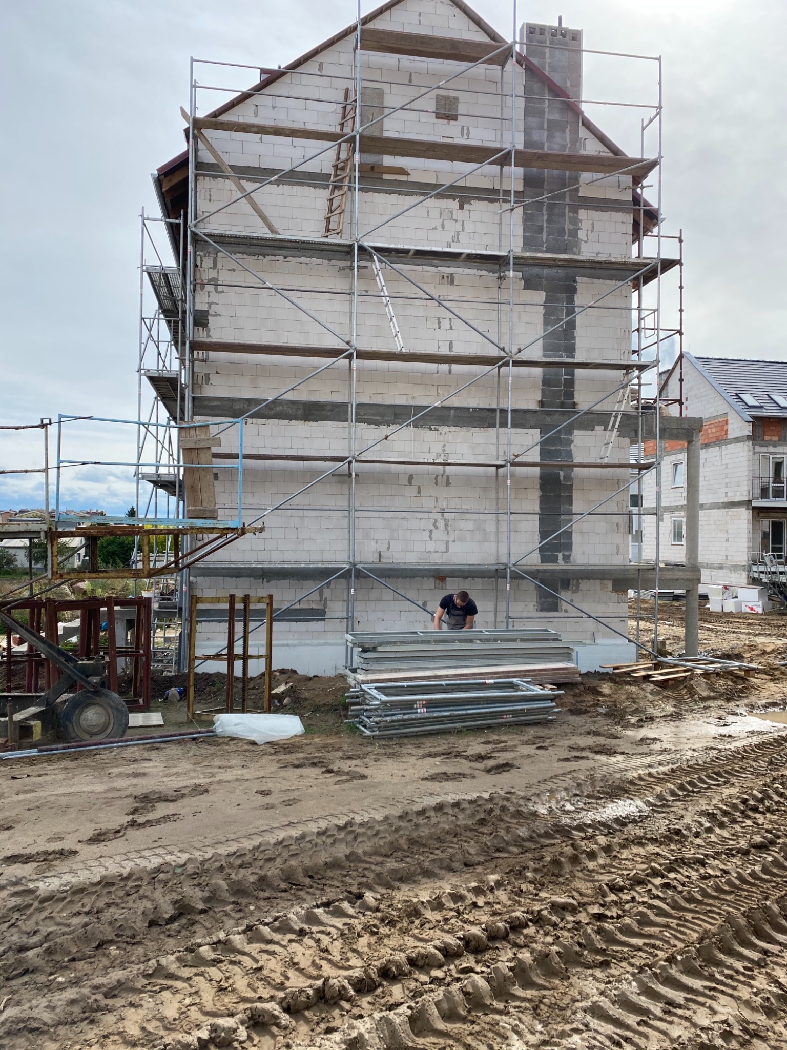Budowa domu jednorodzinnego z białych bloczków, widoczne rusztowania, komin w trakcie budowy, teren wokół rozkopany, pracownik w czarnej koszulce przy ścianie budynku.