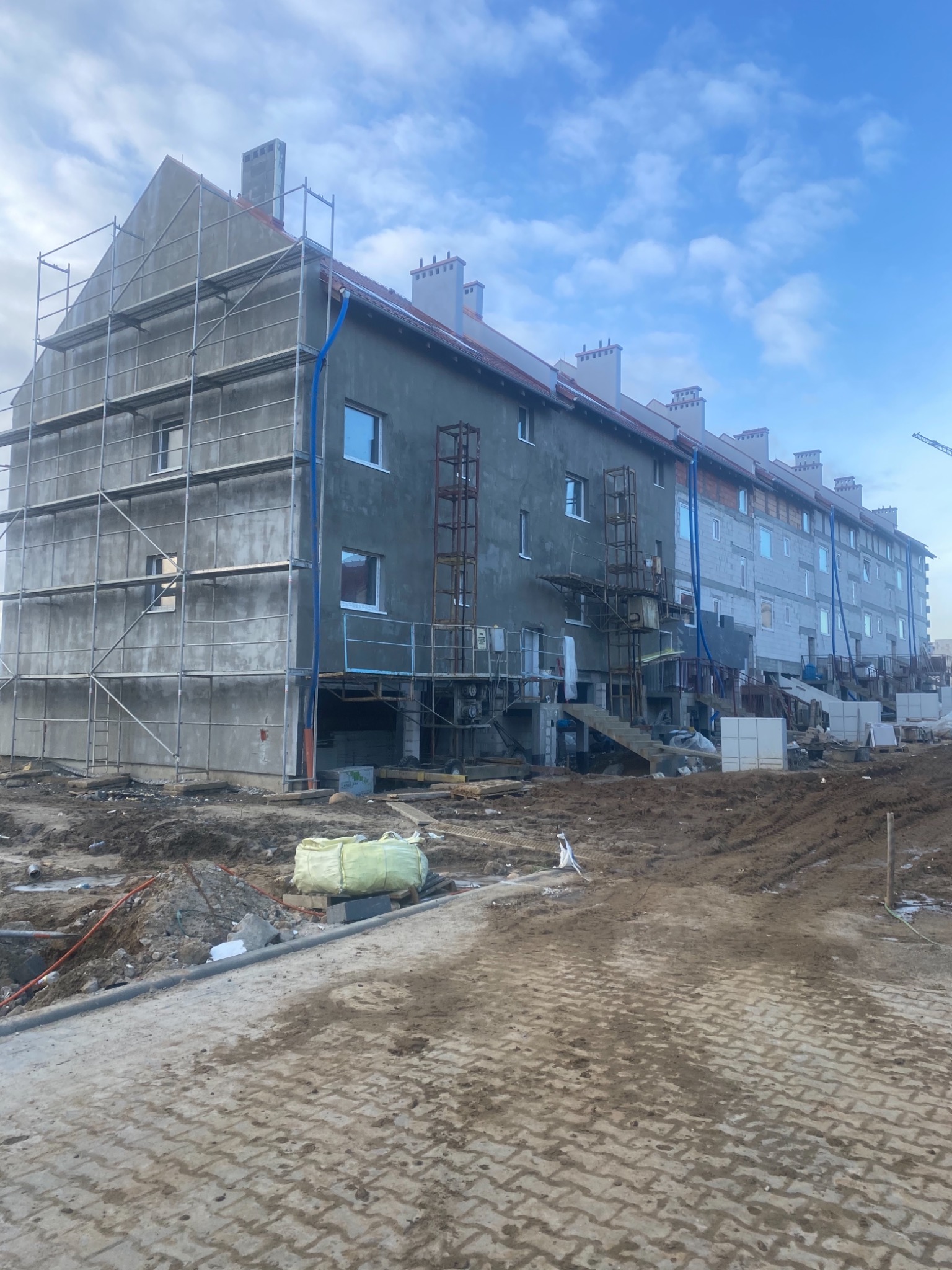 Długi budynek mieszkalny w trakcie prac elewacyjnych; część budynku pokryta szarym tynkiem, pozostała część w trakcie przygotowania; widoczne rusztowania, drabiny, worki z materiałami budowlanymi...