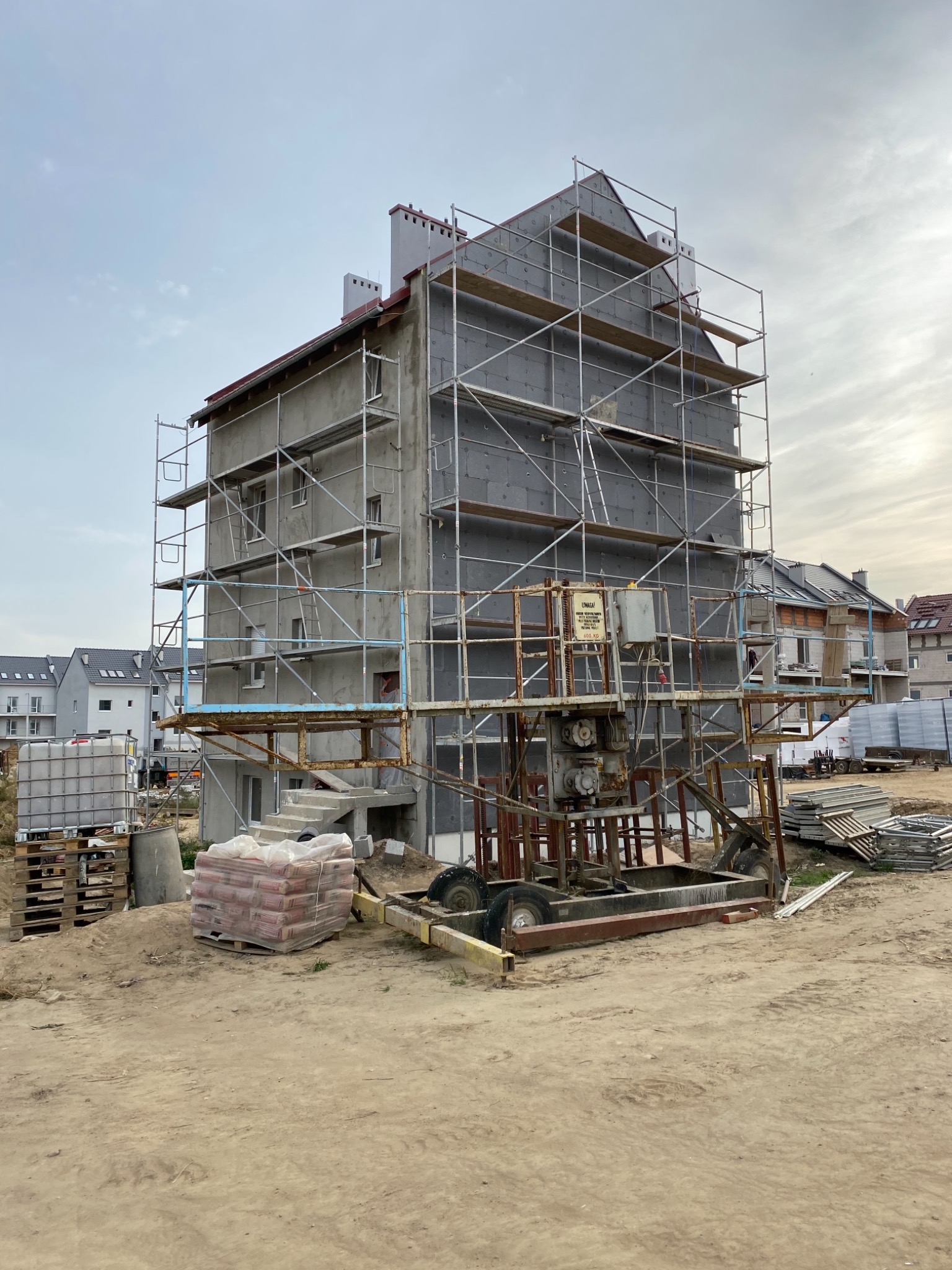 Budynek w trakcie prac ociepleniowych, widoczne rusztowania, płyty izolacyjne na elewacji i maszyna do transportu materiałów budowlanych.