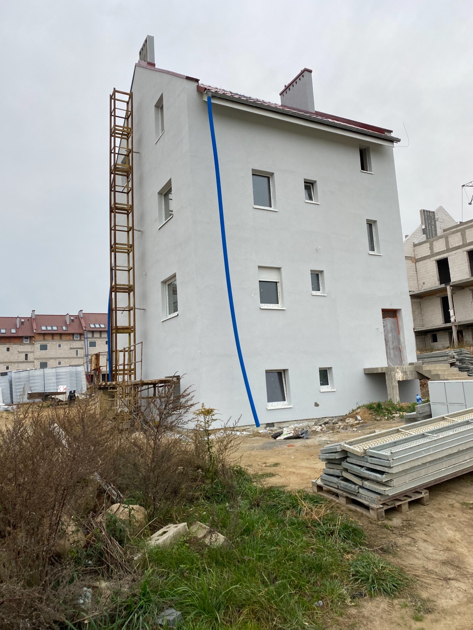 Wysoki, trzypiętrowy budynek po świeżym pomalowaniu na biało, z zamontowaną metalową drabiną rusztowania po lewej stronie i niebieskim przewodem wzdłuż elewacji, na tle placu budowy z innymi...