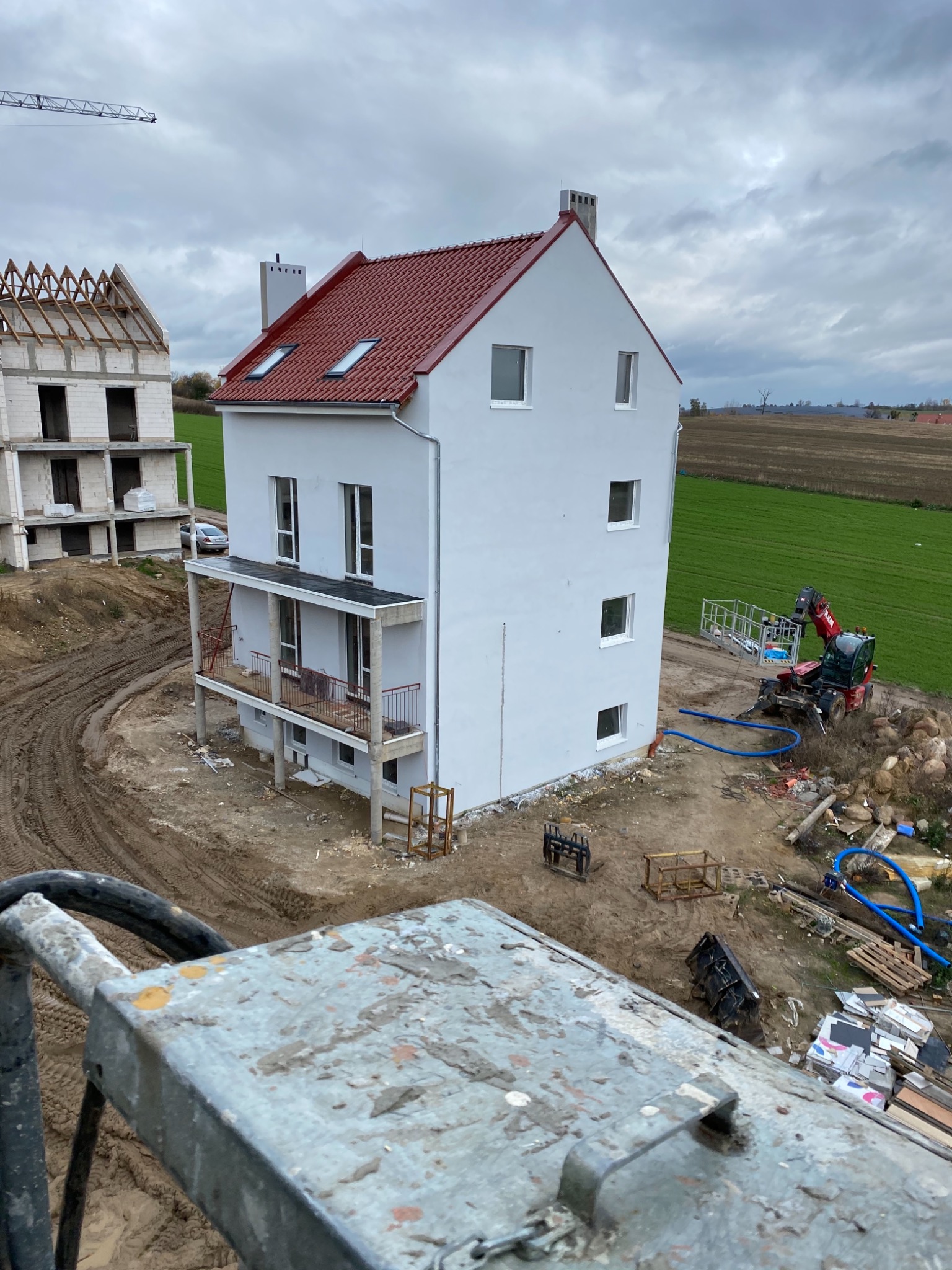 Nowo otynkowany, trzypiętrowy dom z czerwonym dachem, widok z góry, w tle pole i drugi budynek w stanie surowym otwartym, na pierwszym planie elementy rusztowania.