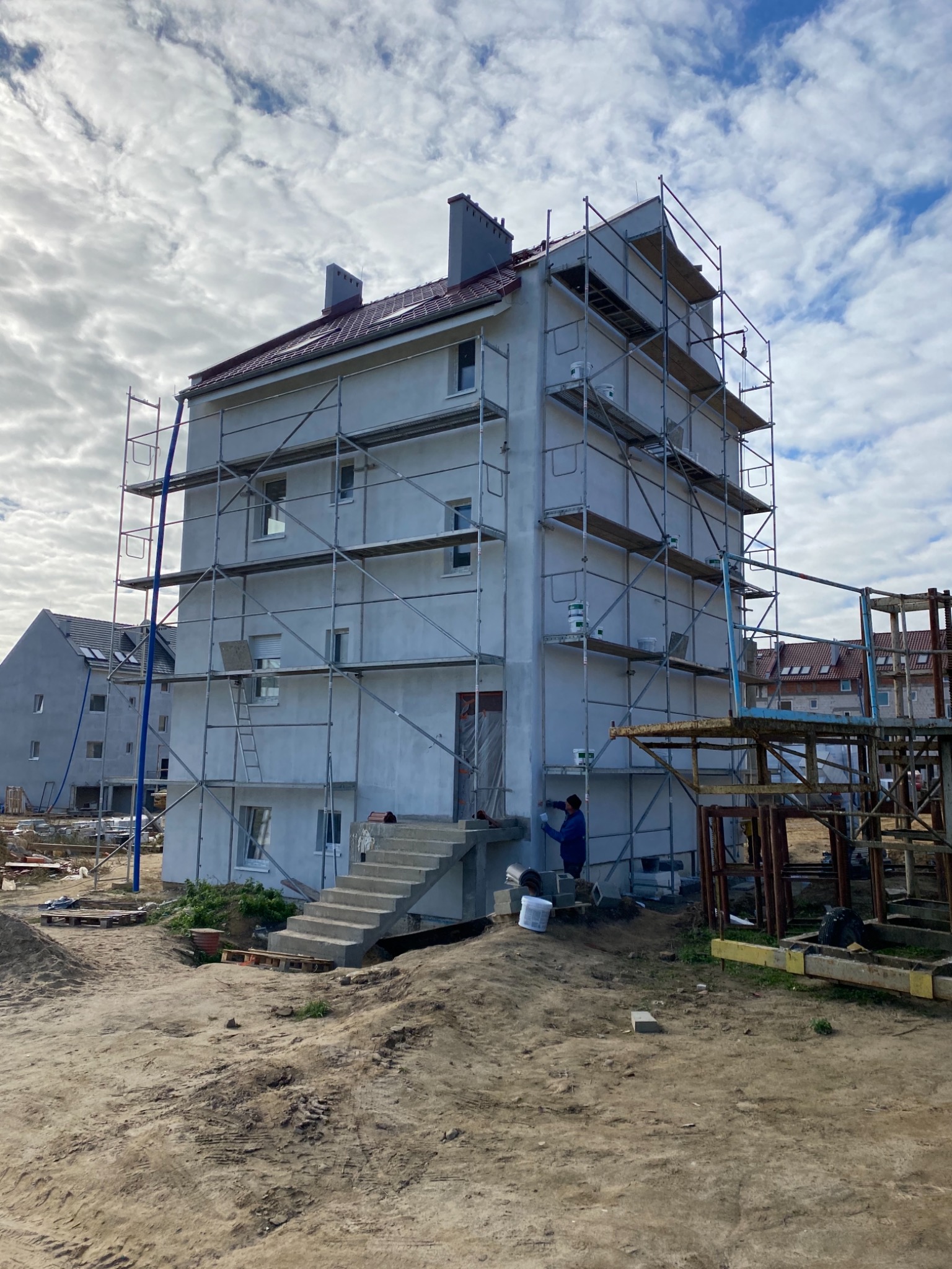 Trzypiętrowy budynek w trakcie prac wykończeniowych elewacji, otoczony rusztowaniami, z widocznym pracownikiem w niebieskiej bluzie przy pracy, na pierwszym planie teren budowy.