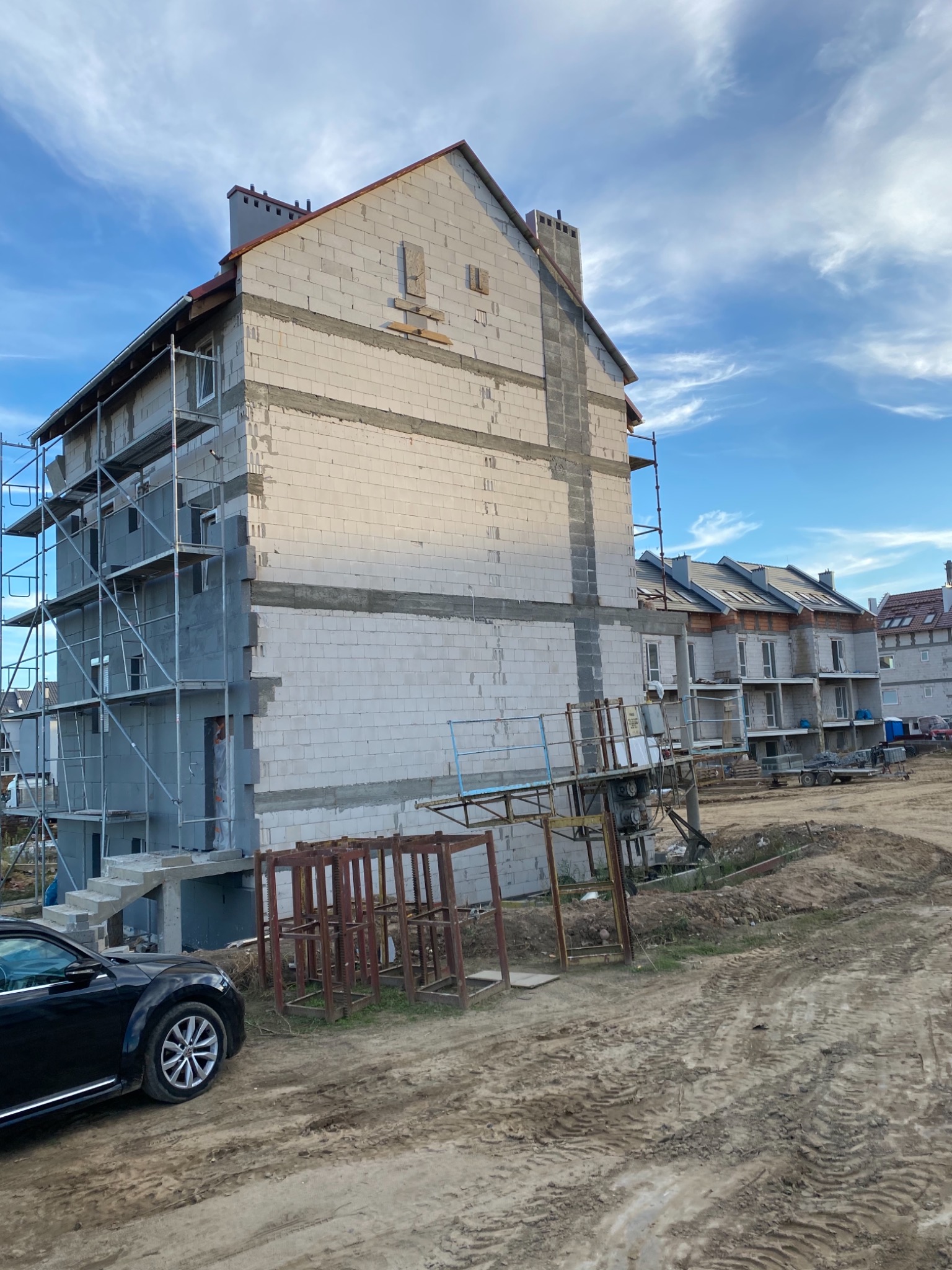 Budynek w trakcie budowy elewacji z widocznymi rusztowaniami, pustakami i elementami konstrukcyjnymi, na tle błękitnego nieba z chmurami.