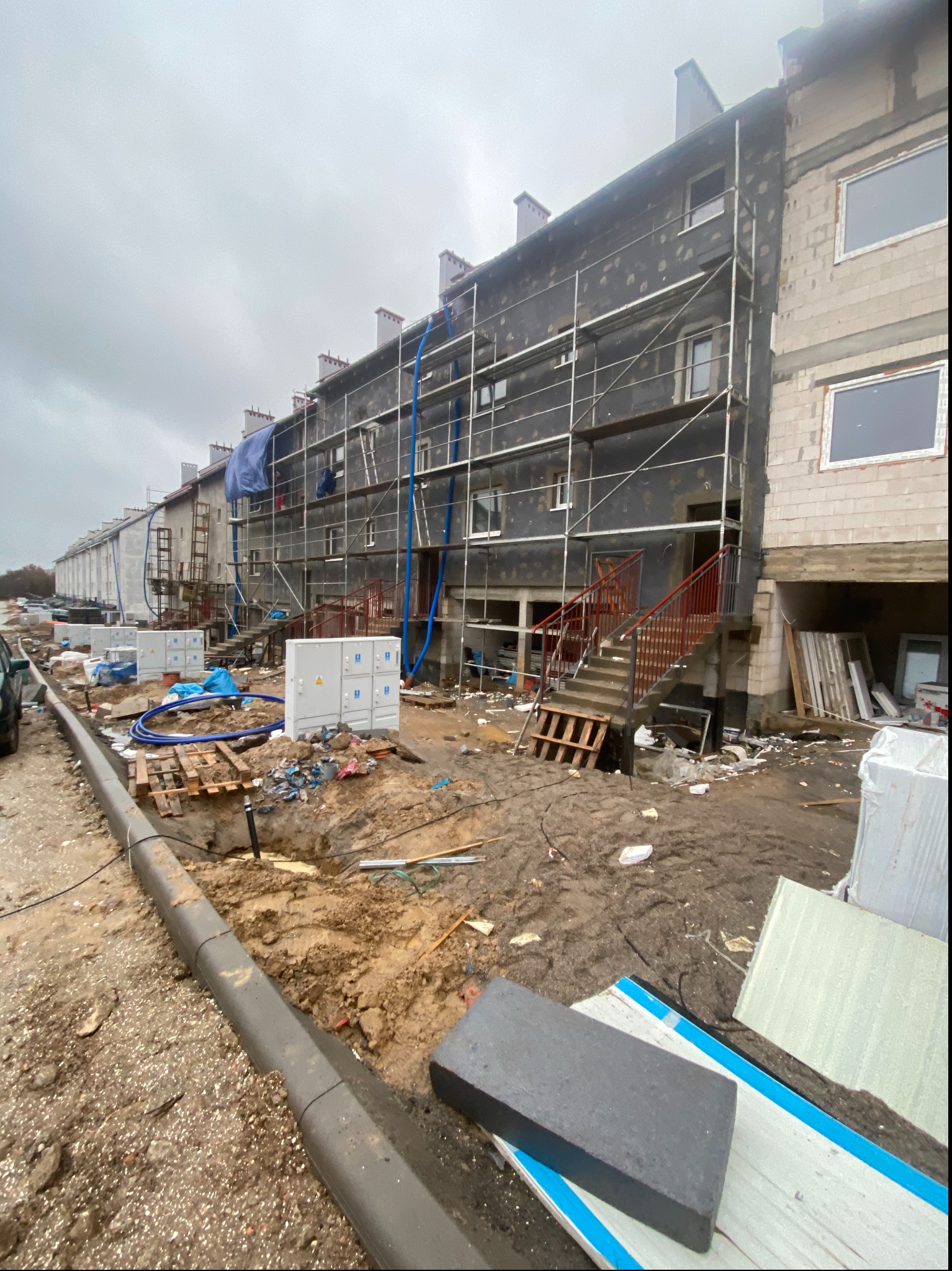 Blok mieszkalny w trakcie prac termoizolacyjnych, widoczne rusztowania, styropian, oraz rozkopany teren przed budynkiem.