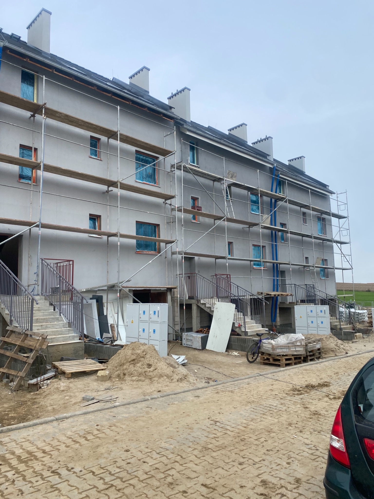 Trzypiętrowy budynek wielorodzinny w trakcie prac elewacyjnych, z rusztowaniami, zabezpieczonymi oknami folią i stertami materiałów budowlanych na placu budowy.