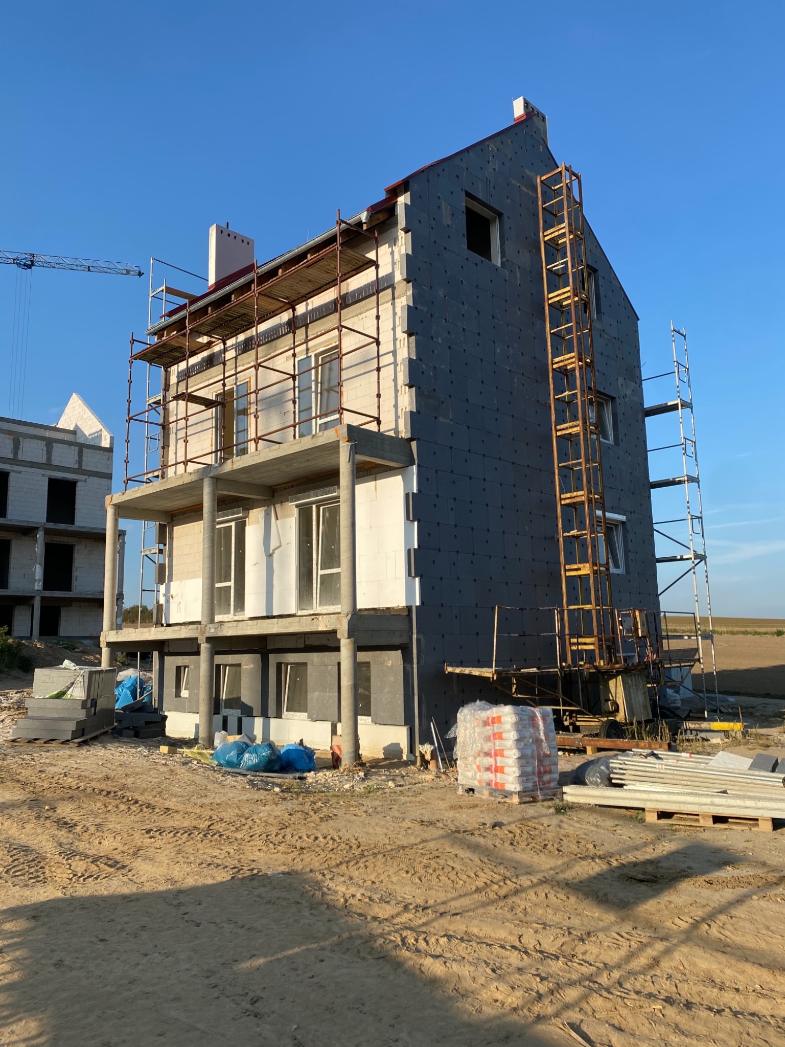 Budynek w trakcie prac elewacyjnych: częściowo ocieplony, z rusztowaniami, widoczne materiały budowlane i elementy konstrukcyjne, niebieskie worki z materiałami leżące na ziemi.