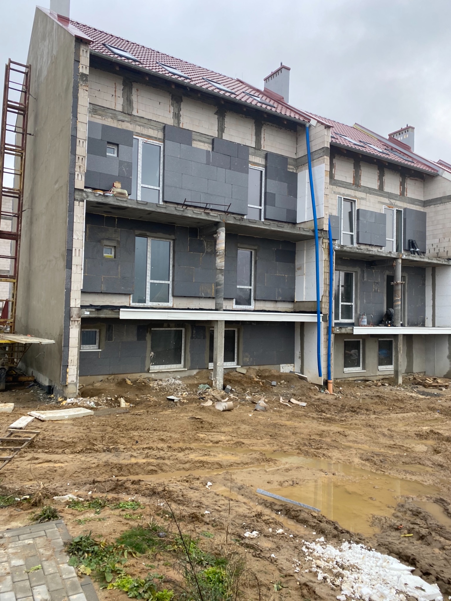 Wielopiętrowy budynek w trakcie ocieplania z widocznymi szarymi płytami izolacyjnymi na elewacji, rusztowanie z boku, teren wokół budynku rozkopany i błotnisty.