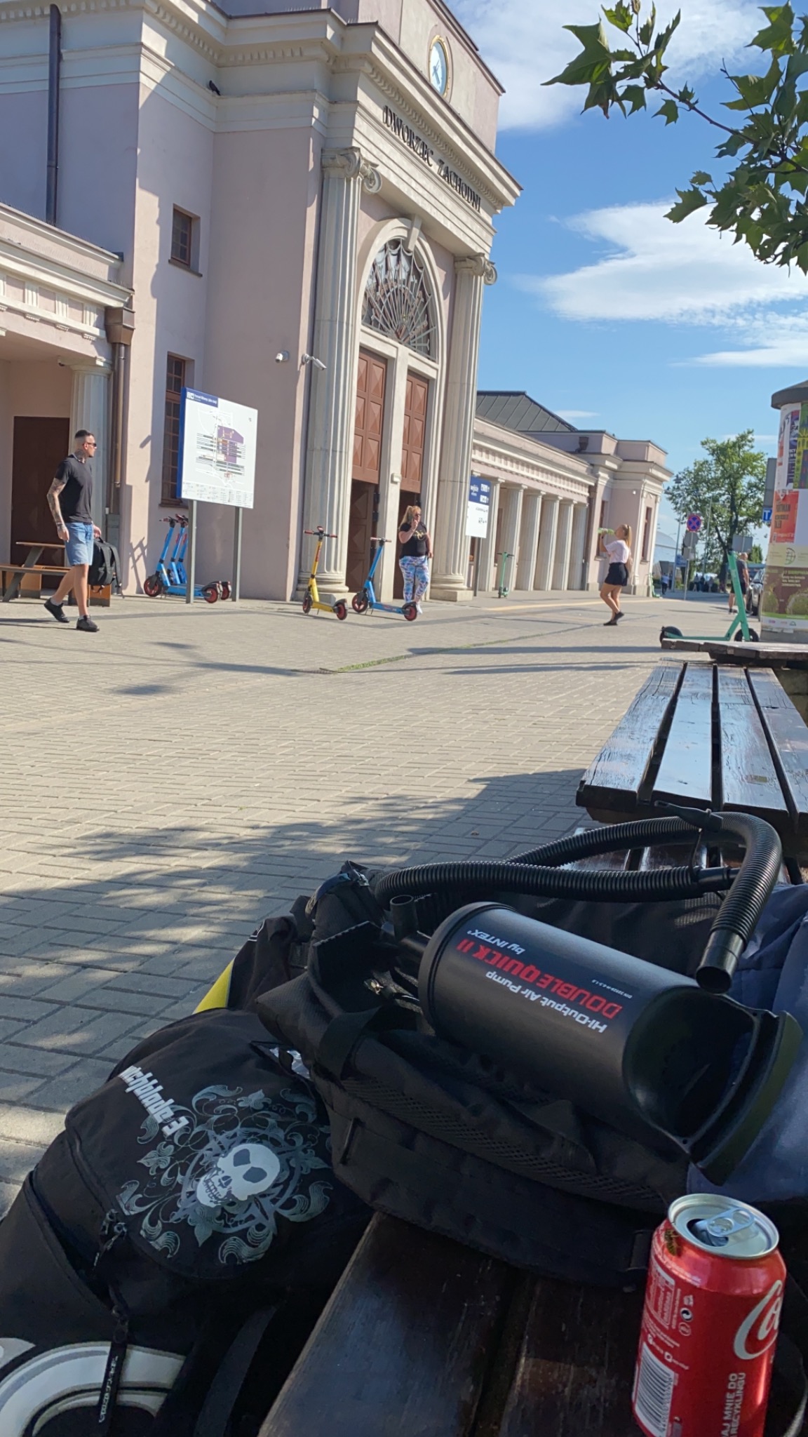 Widok na Dworzec Zachodni z poziomu ławki, na której leżą plecaki, pompka Intex Double Quick II i pusta puszka Coca-Coli.