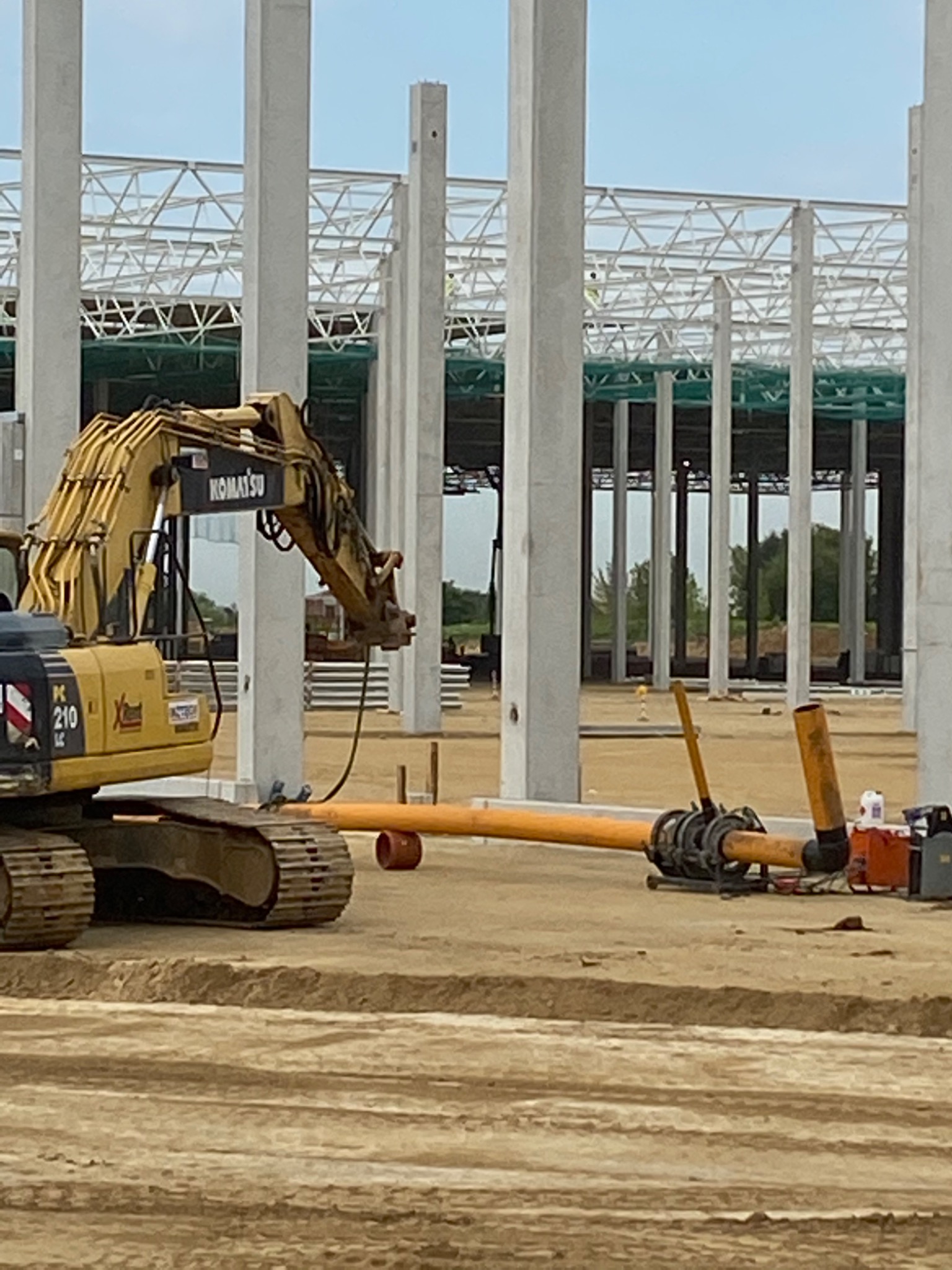 Żółta koparka Komatsu na placu budowy hali magazynowej, rury kanalizacyjne w wykopie ziemnym.