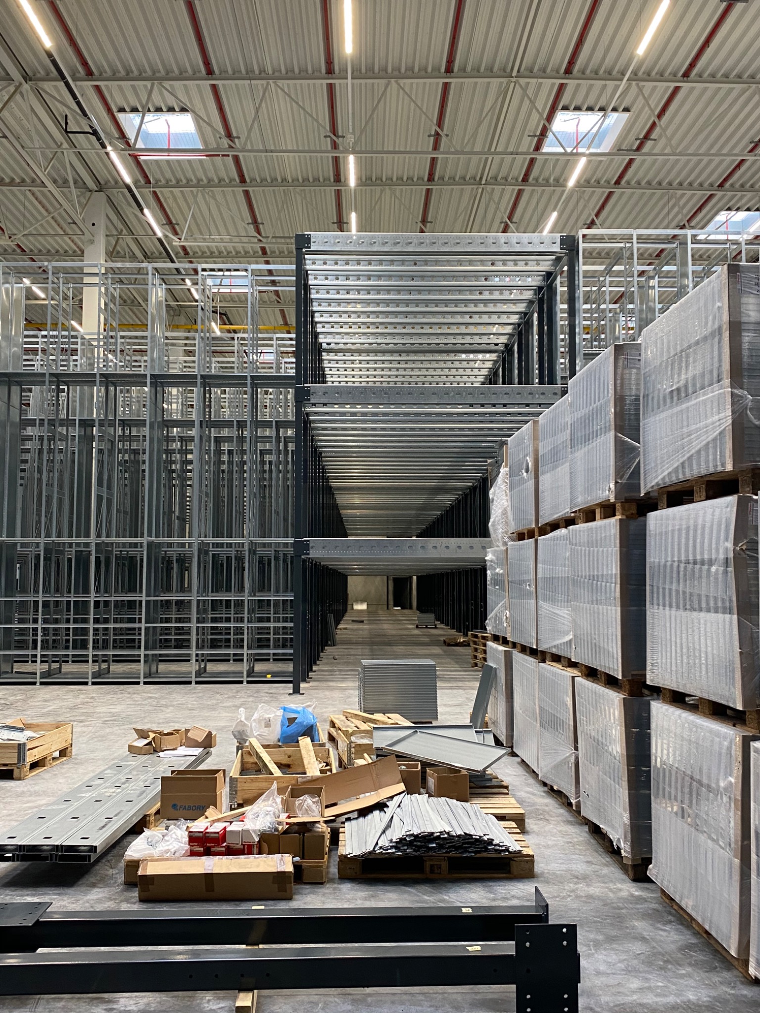 Wnętrze hali magazynowej w trakcie budowy regałów wysokiego składowania, widoczne elementy konstrukcyjne, palety z towarem, kartony i materiały montażowe.