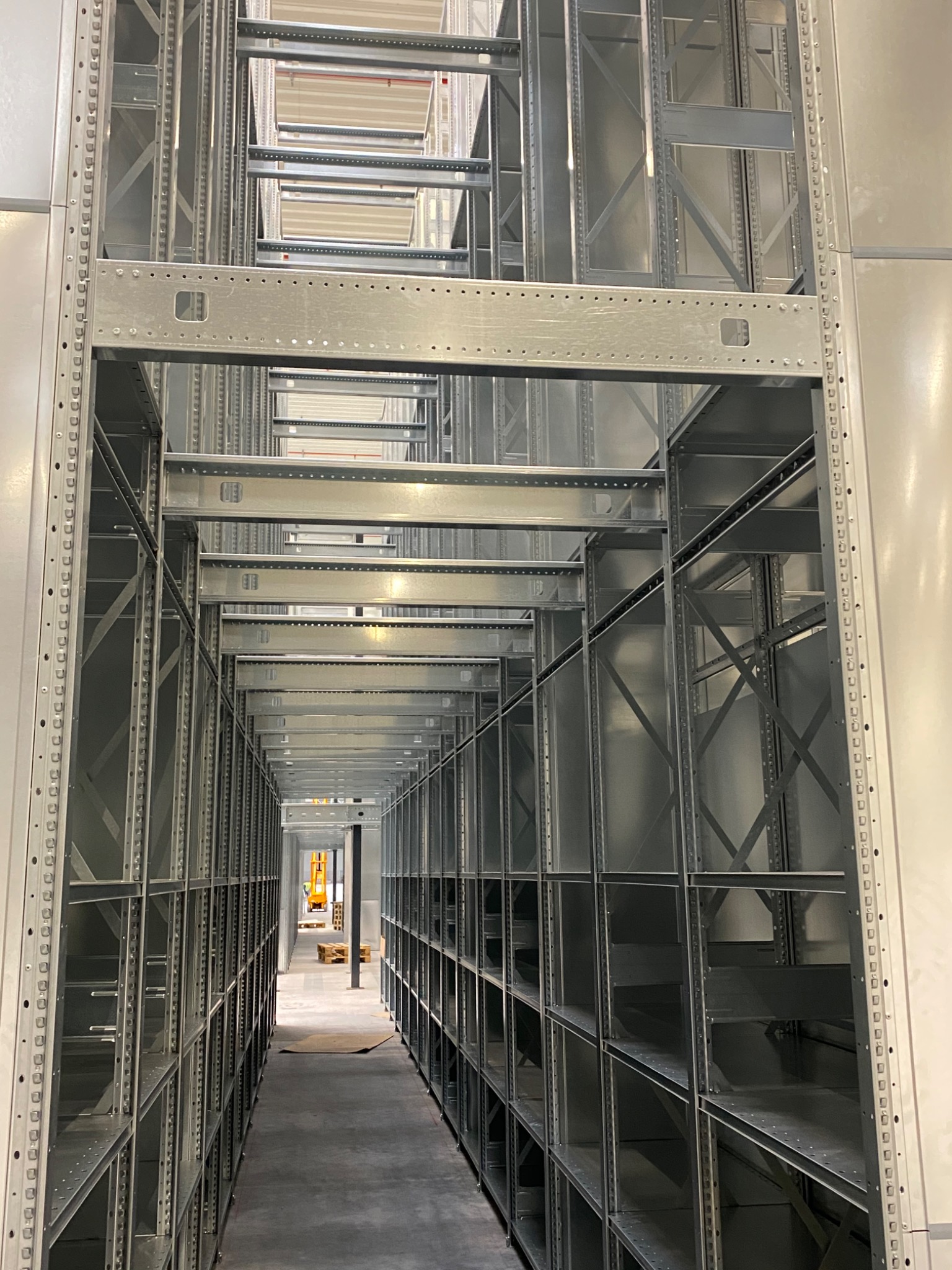 Pusty korytarz między wysokimi, metalowymi regałami magazynowymi, perspektywa z dołu, w oddali widoczny wózek widłowy.