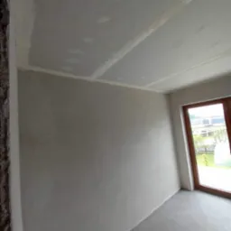 Wnętrze pokoju w trakcie remontu, ściany pokryte szarą gładzią, widoczne łączenia płyt kartonowo-gipsowych na suficie, okno z drewnianą ramą.