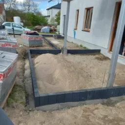 Przygotowanie terenu pod taras: ułożone obrzeża z kostki brukowej, wypełnione piaskiem, palety z kostką brukową w tle, widoczny fragment elewacji budynku.