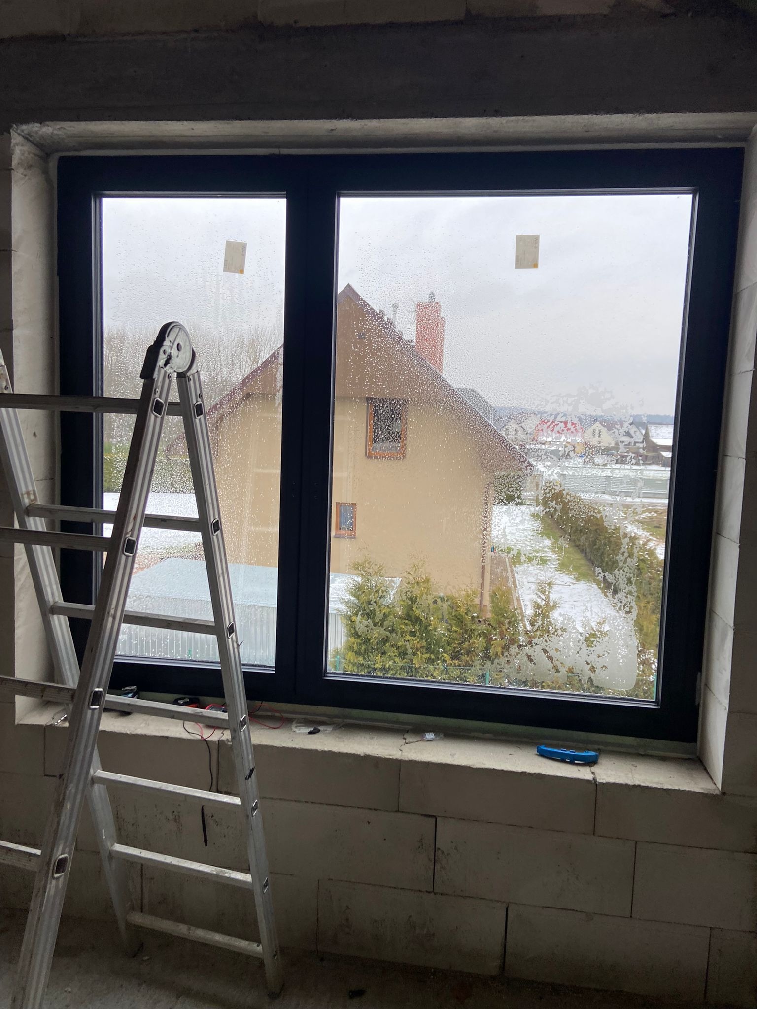 Nowe, czarne okno zamontowane w surowym stanie, widoczne ślady wilgoci na szybach, drabina oparta o ścianę, widok na dom i częściowo ośnieżony ogród za oknem.