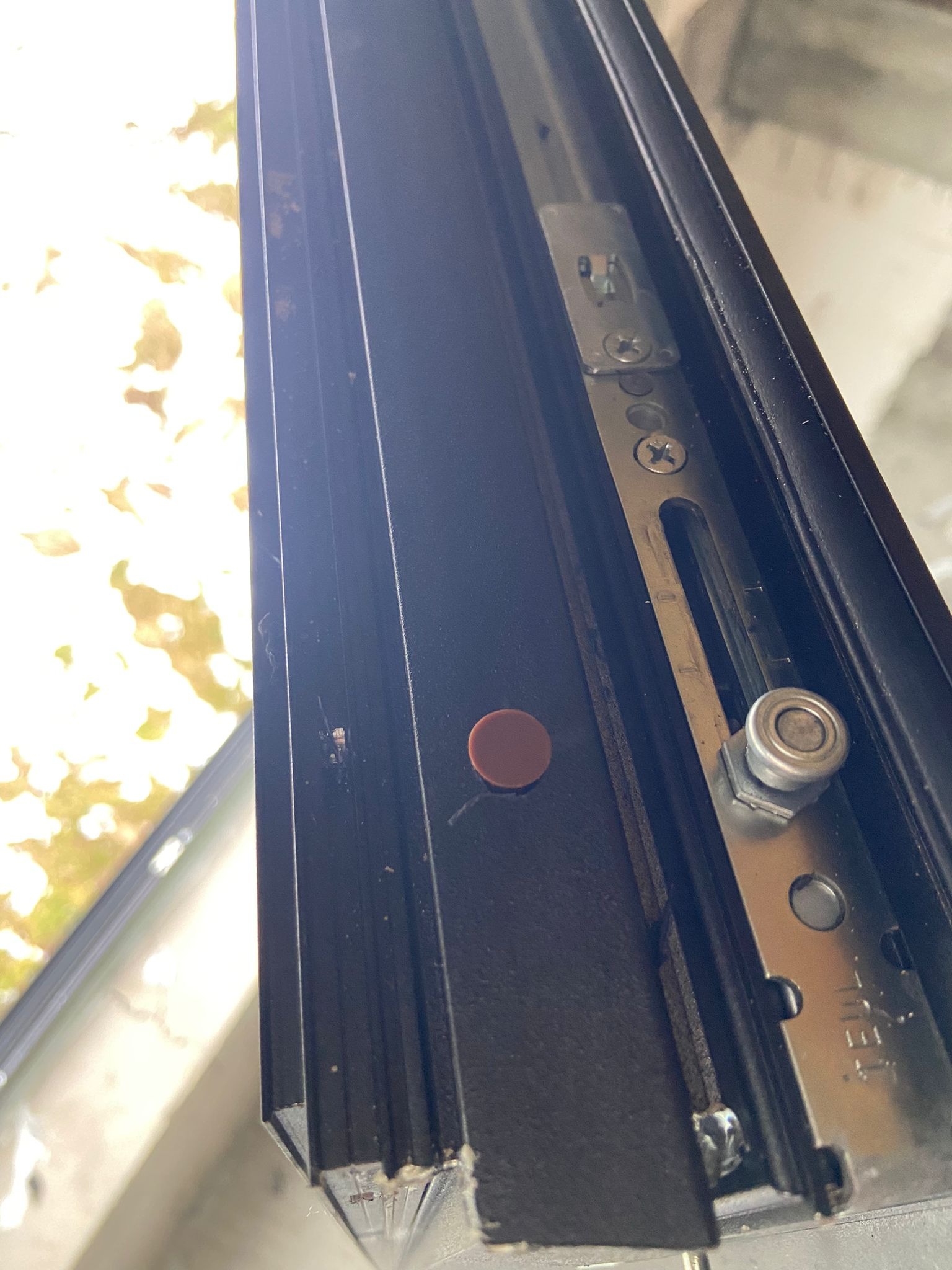 Szczegół mechanizmu okiennego z widocznymi śrubami regulacyjnymi i rolką, czarna rama okienna na tle rozmytej zieleni.