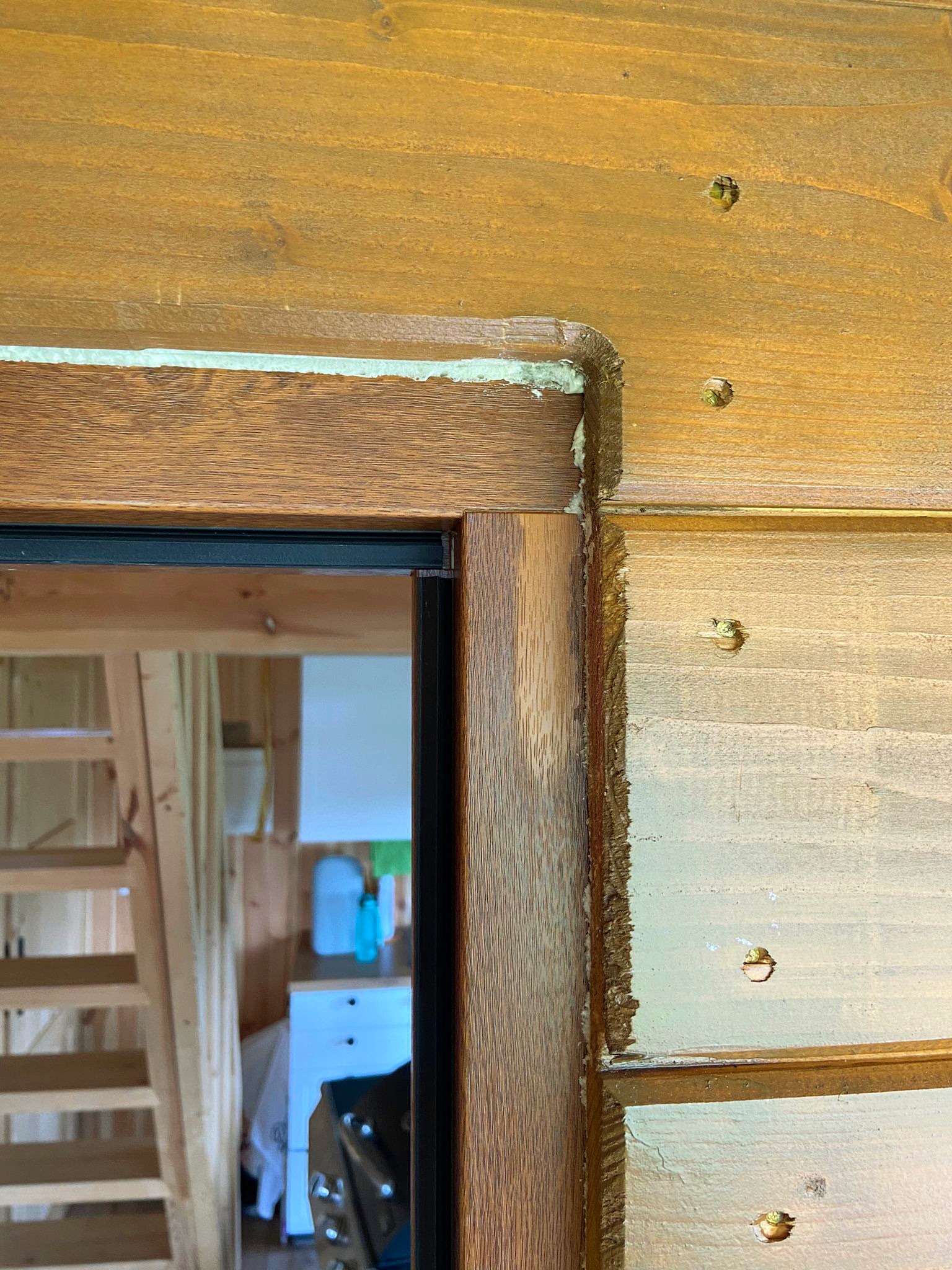 Fragment montażu drewnianej ościeżnicy drzwiowej z widocznymi śladami uszczelniania i wkrętami, w tle widoczna drewniana konstrukcja schodów.